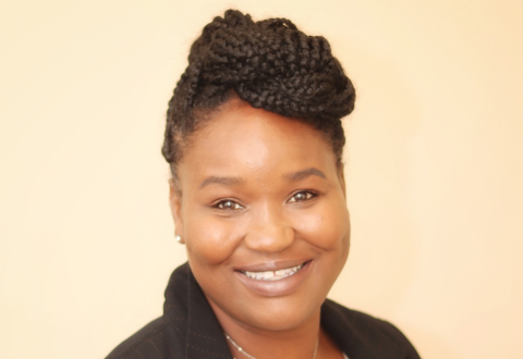 Headshot of a black women with braided hair in a top knot. She is standing in front of a cream background, smiling, wearing a black blazer.
